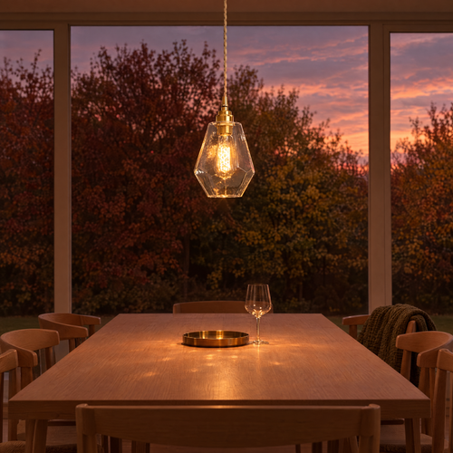 Aurum faceted glass pendant light hanging low over a pale oak dining table, autumn trees and a dusk sky through full-height glazing
