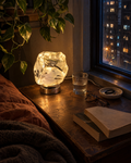 Glacia rock-cut glass table lamp in clear silver, lit on a bedside table beside a rainy city window at night