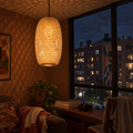 Tessera woven bamboo pendant lit at night in a city apartment, its lattice casting a lacework of light across the wall and ceiling