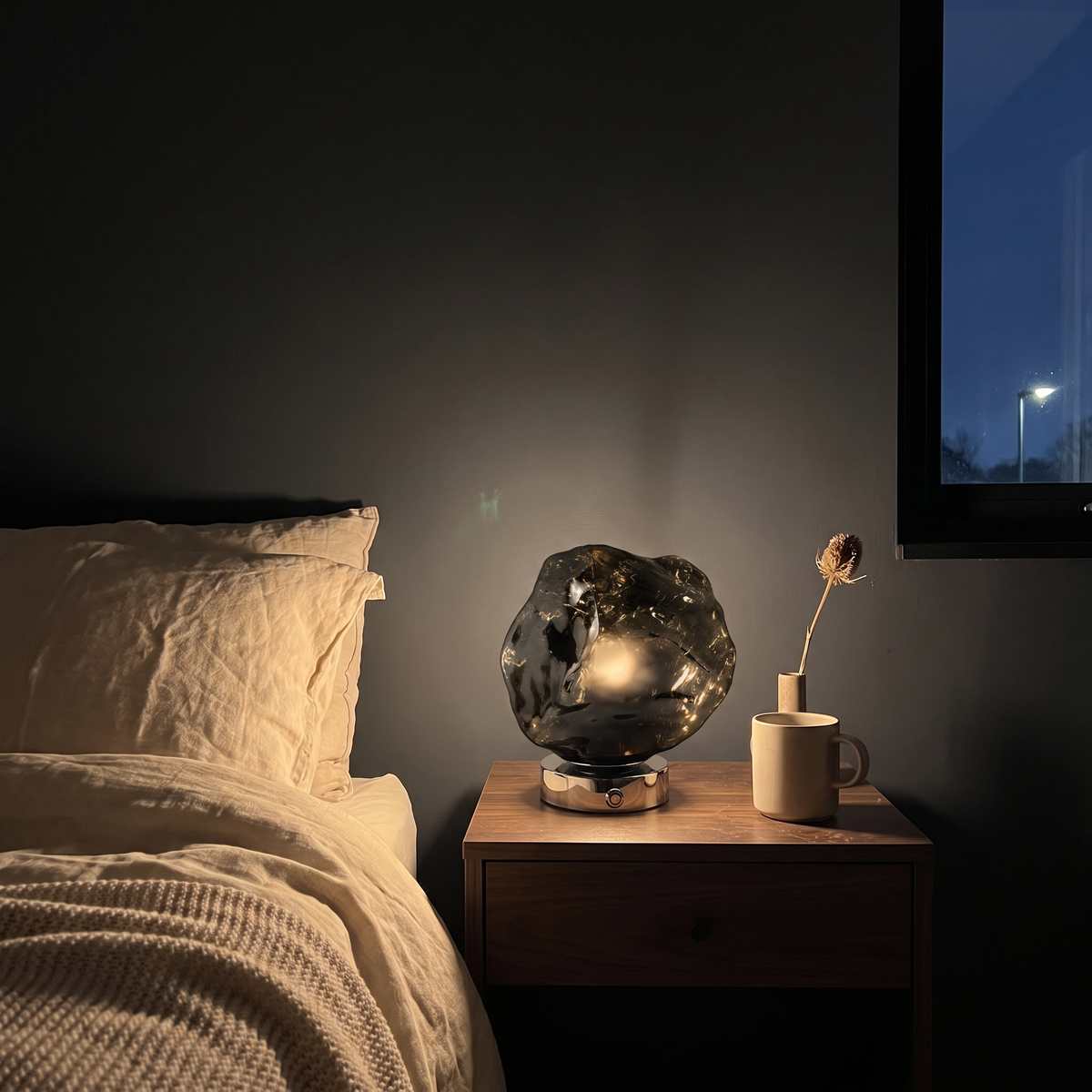 Glacia rock-cut glass table lamp in smoke gray, lit on a walnut nightstand in a dark bedroom