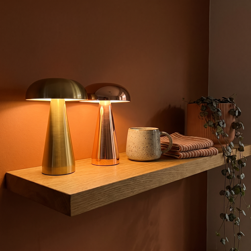 Morel table lamp in brushed brass and rose gold side by side on an oak shelf, both lit